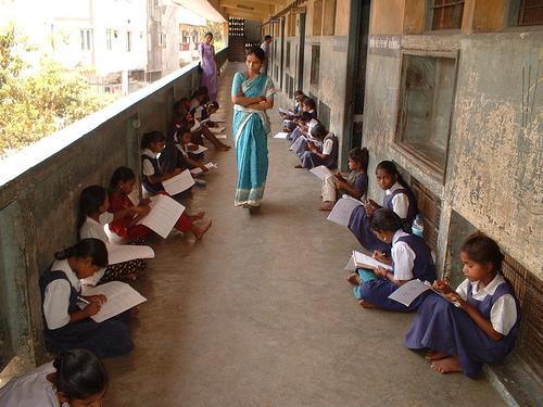 Uniformed students sit on school porch as teacher in sari watches