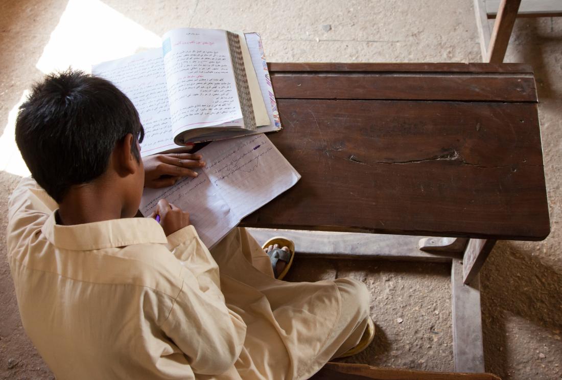 Young boy sitting at desk reads a textbook and writes in a notebook.