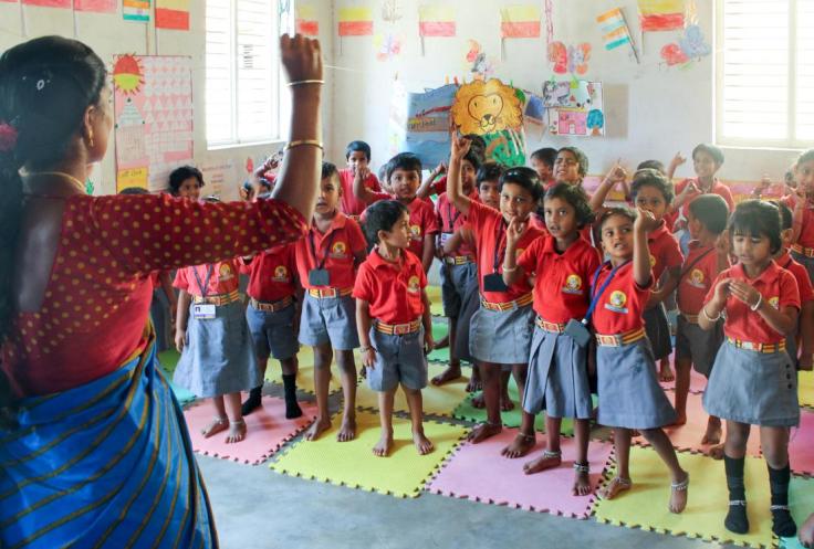 Children stand with their hands raised in a kindergarten classroom.