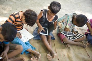 Children sit on the ground, playing on tablets
