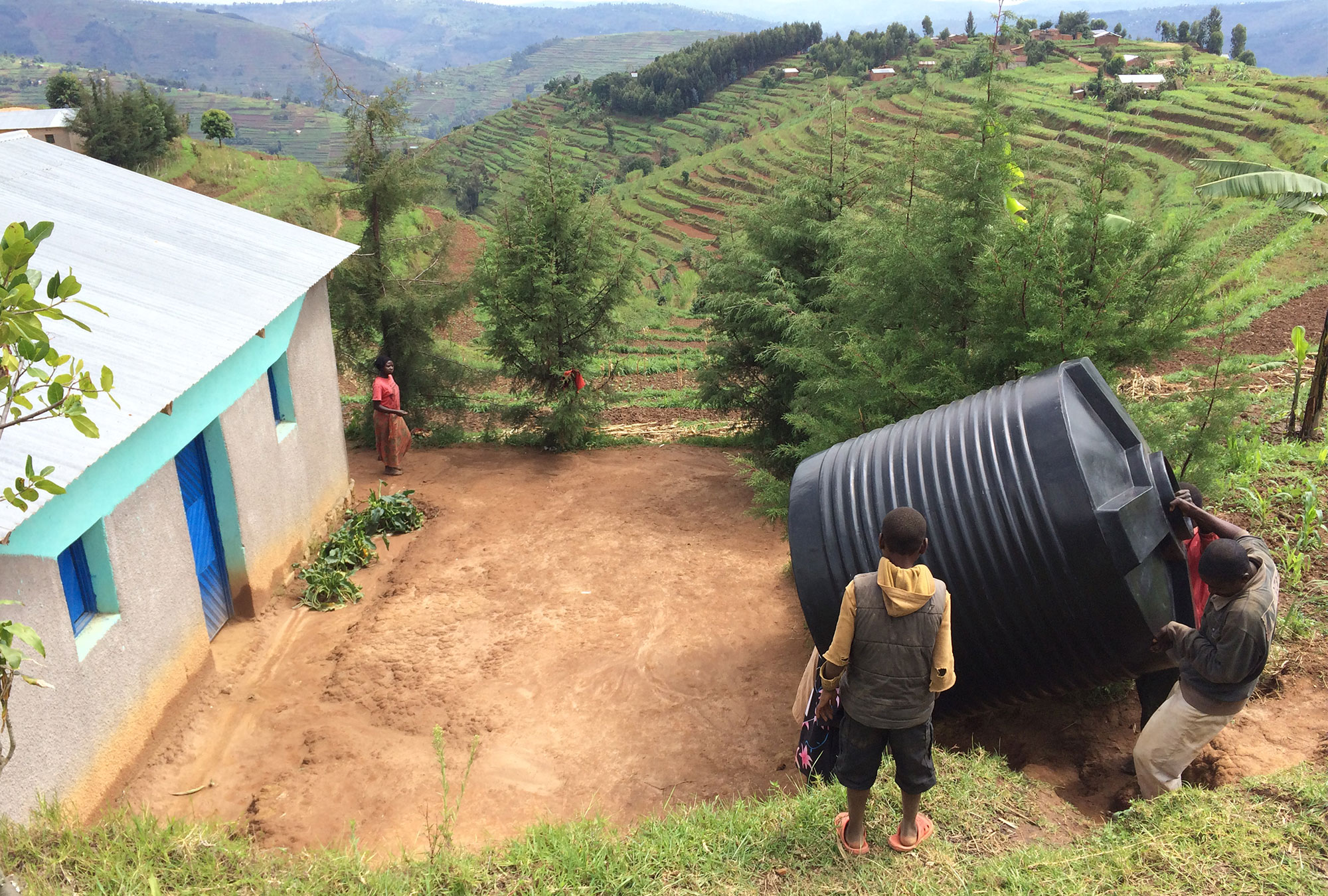 People carry large water tank up a hill in terraced agricultural landscape