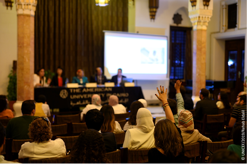 A table of panelists face an audience, several of whom are raising hands
