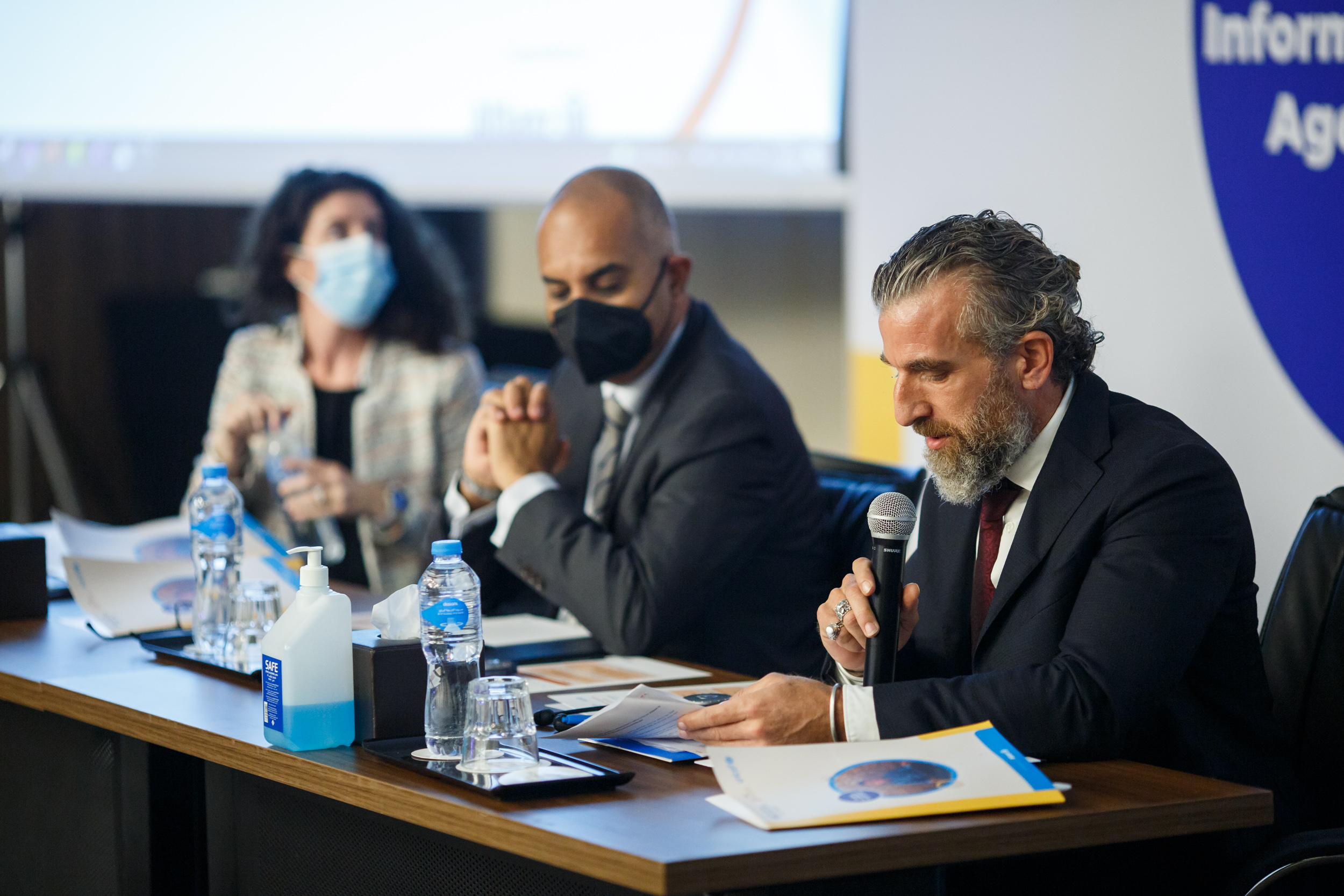 Man speaking into the microphone at table with two other individuals behind him