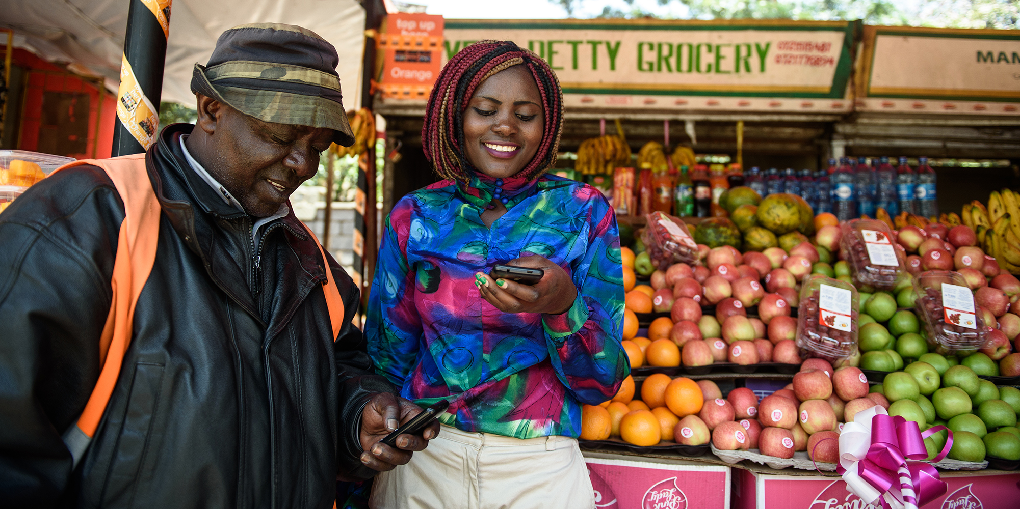 Smiling man looking at a cell phone a woman is holding in front of a fruit stand