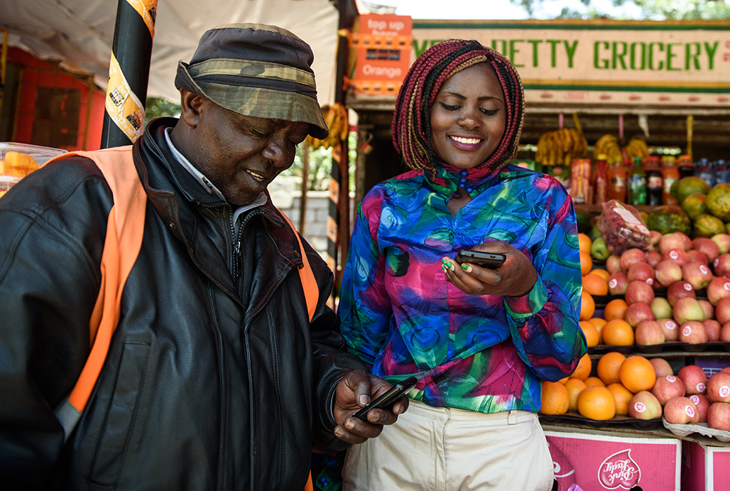 Smiling man looking at a cell phone a woman is holding in front of a fruit stand