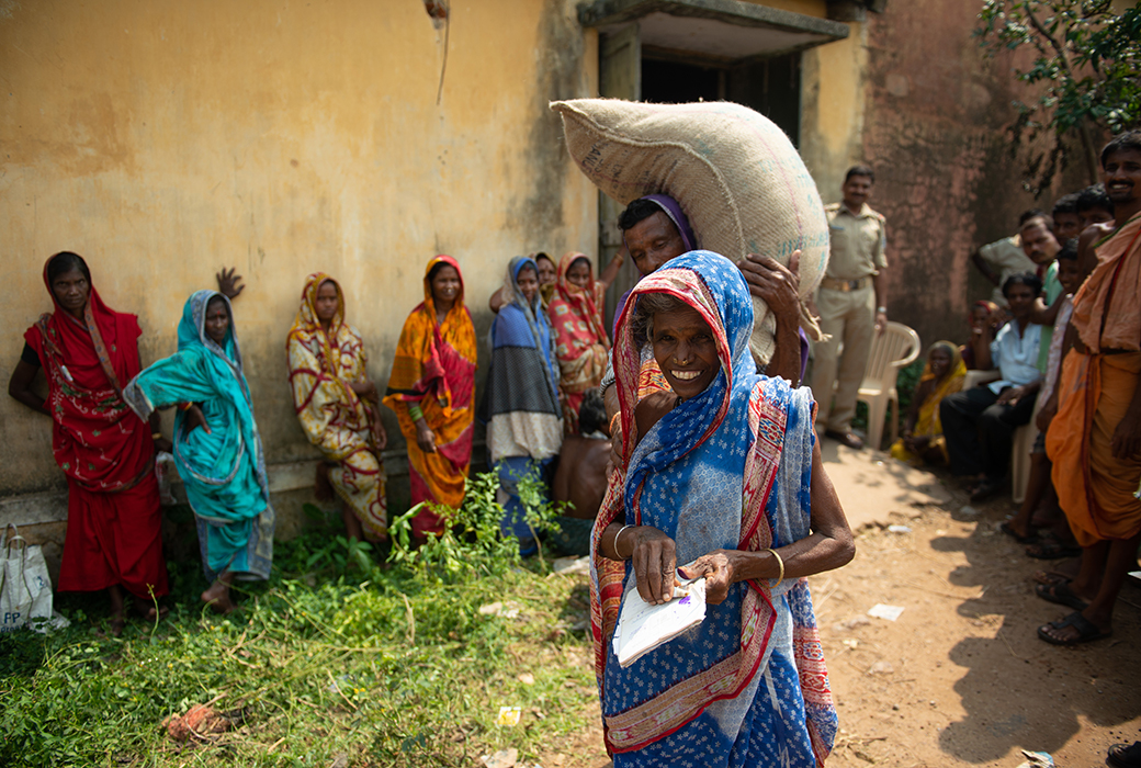 Smiling woman standing in front of a man carrying a bag of rice on his shoulder. Several women and men stand behind them.