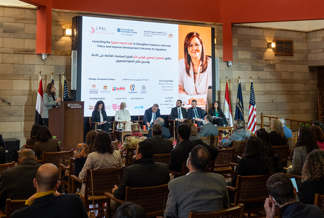 Woman stands at podium speaking alongside six other speakers, talking to a large audience