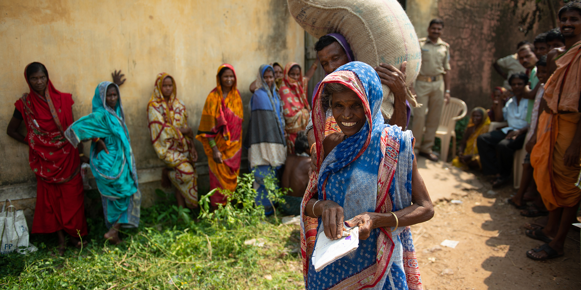 Smiling woman standing in front of a man carrying a bag of rice on his shoulder. Several women and men stand behind them.