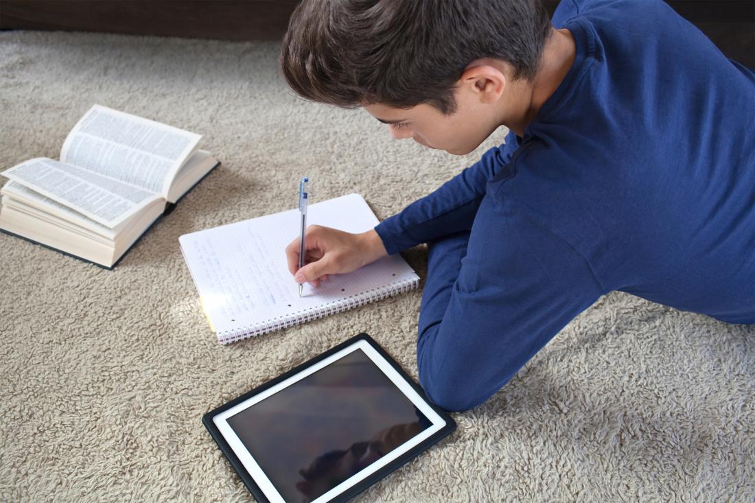 A boy lays on the ground writing in a notebook with a tablet and book next to him. 