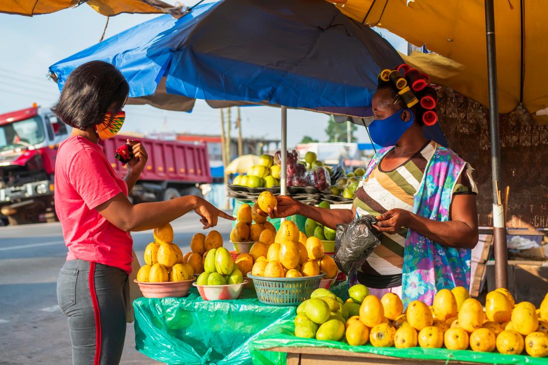 A woman sells fruit to another woman at a market stand while both are wearing face masks over their mouth and nose.