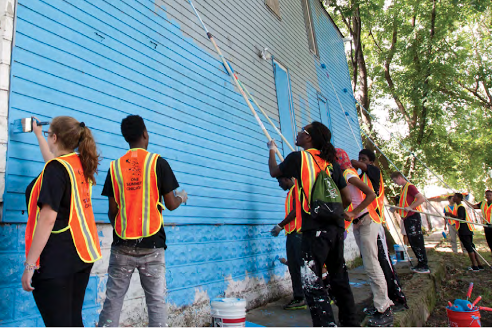 A group of teenagers working outside, wearing safety vests that say "One Summer Chicago" on the back, paint a brick wall blue.