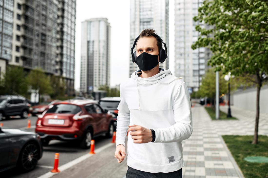 A young man wearing a fit bit, black surgical mask, and white hoodie runs on the sidewalk with cars and buildings in the background.