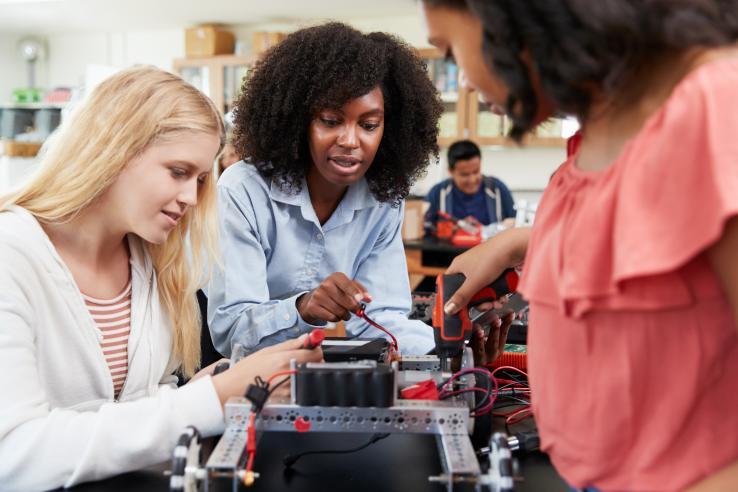 Three young women gathered around a lab bench build a robot together.