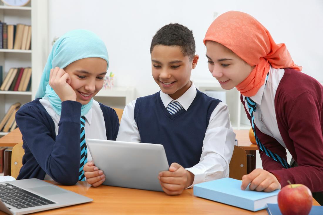 Three school children sitting at a table looking at one tablet.