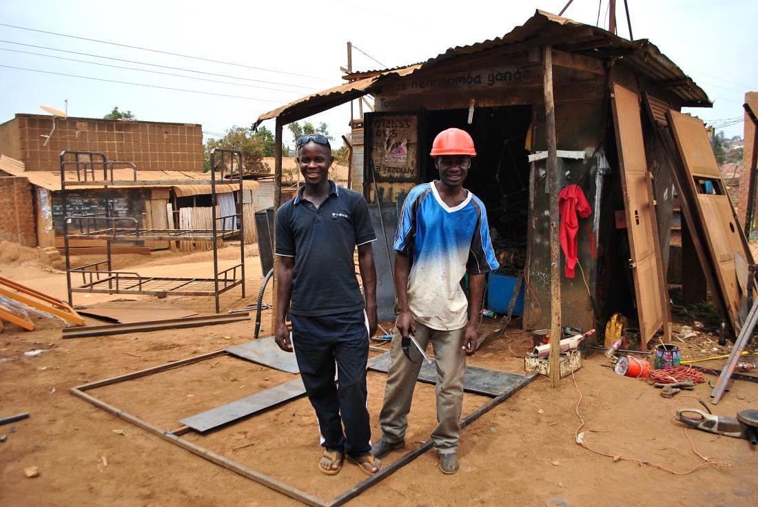 Two men standing outdoors in front of a workshop in Uganda