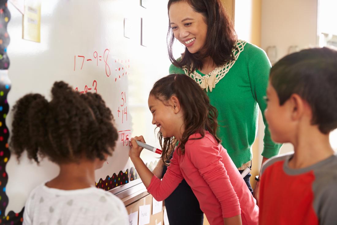 A student does math on a whiteboard while a teacher looks on smiling and classmates gather around.