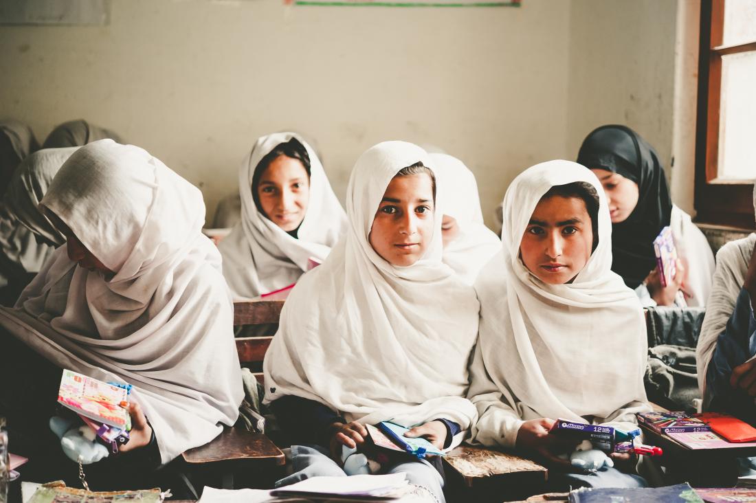 Three girls wearing hijabs sit in a classroom holding books and other learning materials
