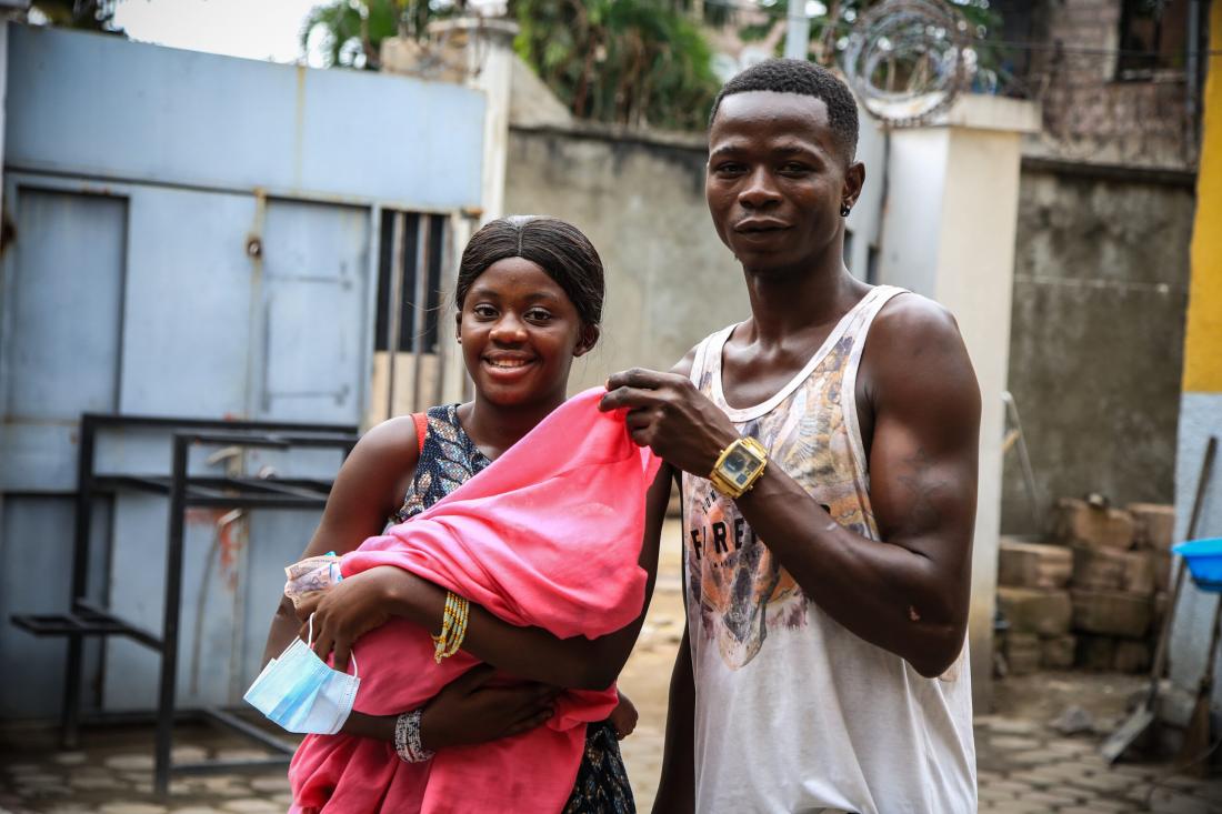 A couple standing looking at the camera carrying a baby.