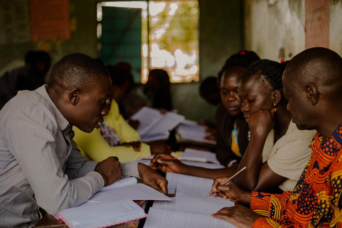 Adults seated in a dim classroom concentrate on notebooks and worksheets.