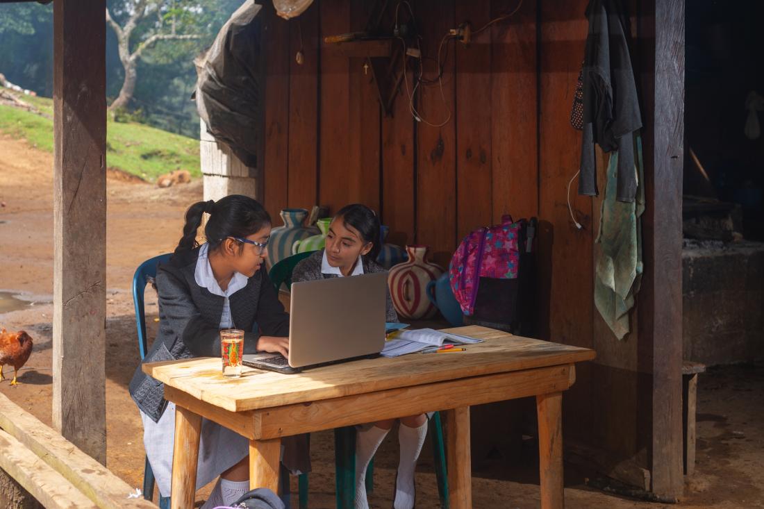 Two girls talk to each other while looking at something on a laptop.