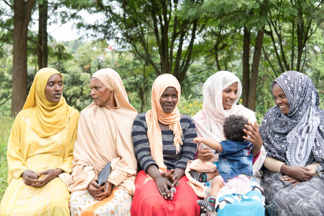 Women sit in a row outside, talking to each other for J-PAL Africa Evaluation Summary
