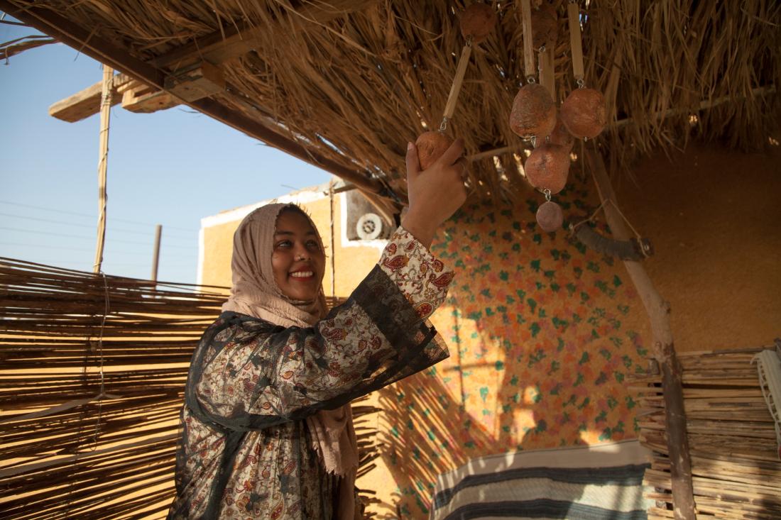 Female youth with wearing headscarf reaches for handmade decorations under a thatched roof in Egypt.