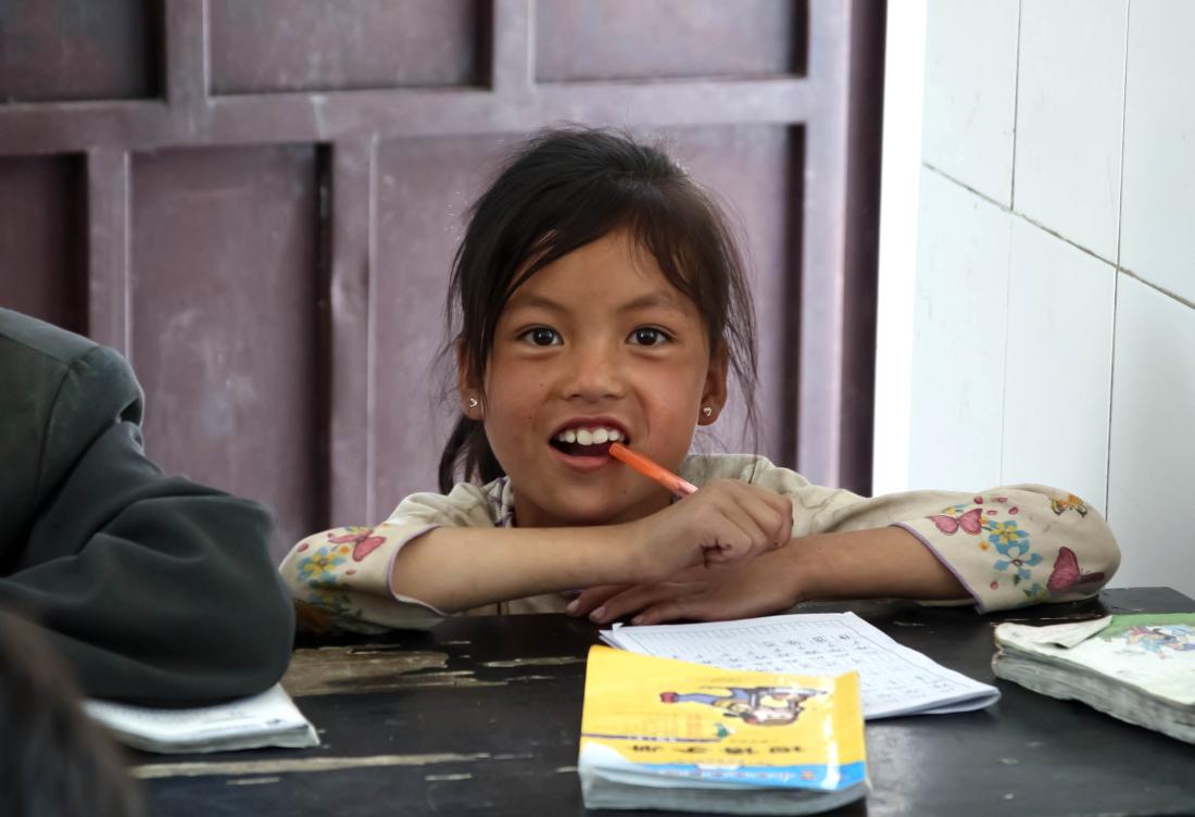 A young Chinese girl sits at desk with pen in mouth and educational workbooks in front of her