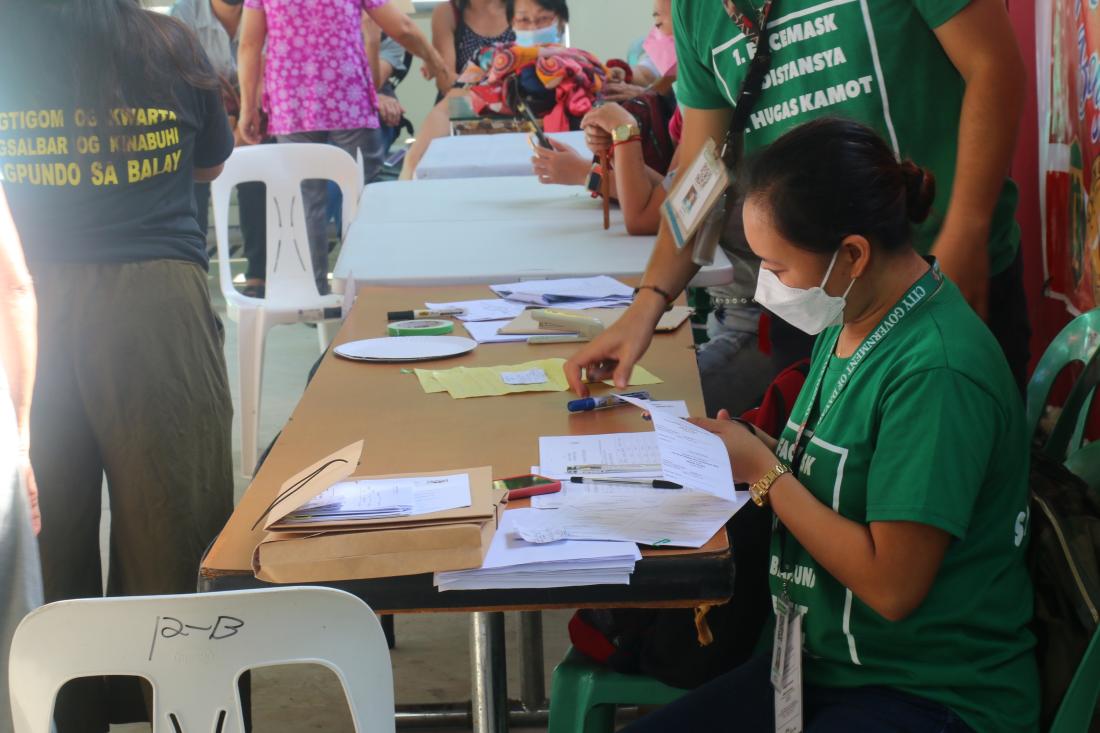 Community development work with masked woman in green shirt sitting at table cutting out piece of paper with other workers in the background