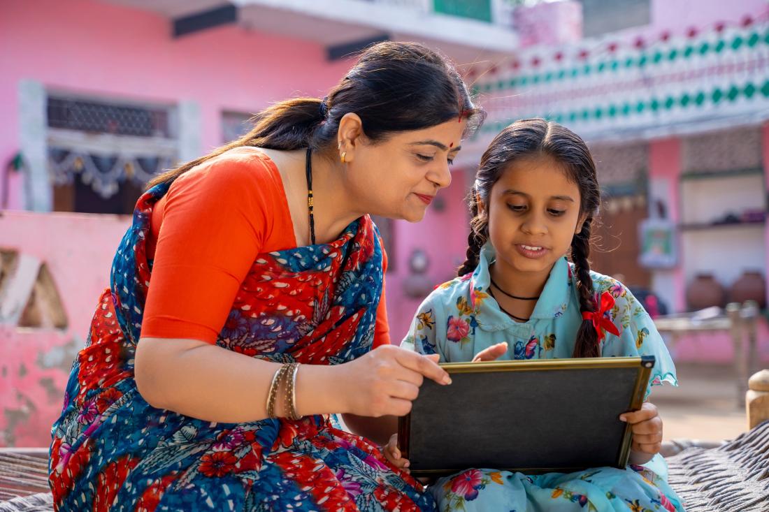 A woman tutoring a young student with braids through a lesson on a tablet at her home in Urban India.