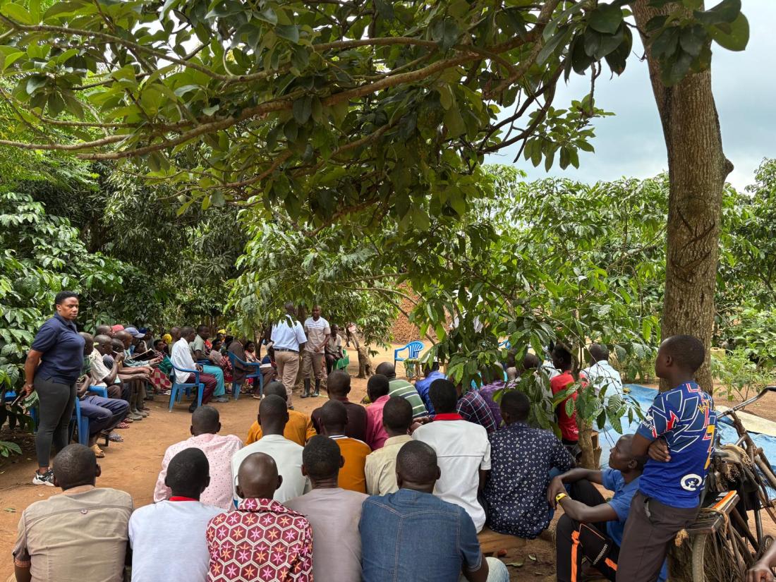 A group of villagers sits in plastic chairs while two men stand and demonstrate agricultural techniques.