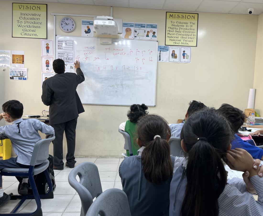 A teacher stands with his back to a classroom of students. The teacher is teaching using the Iqra curriculum.
