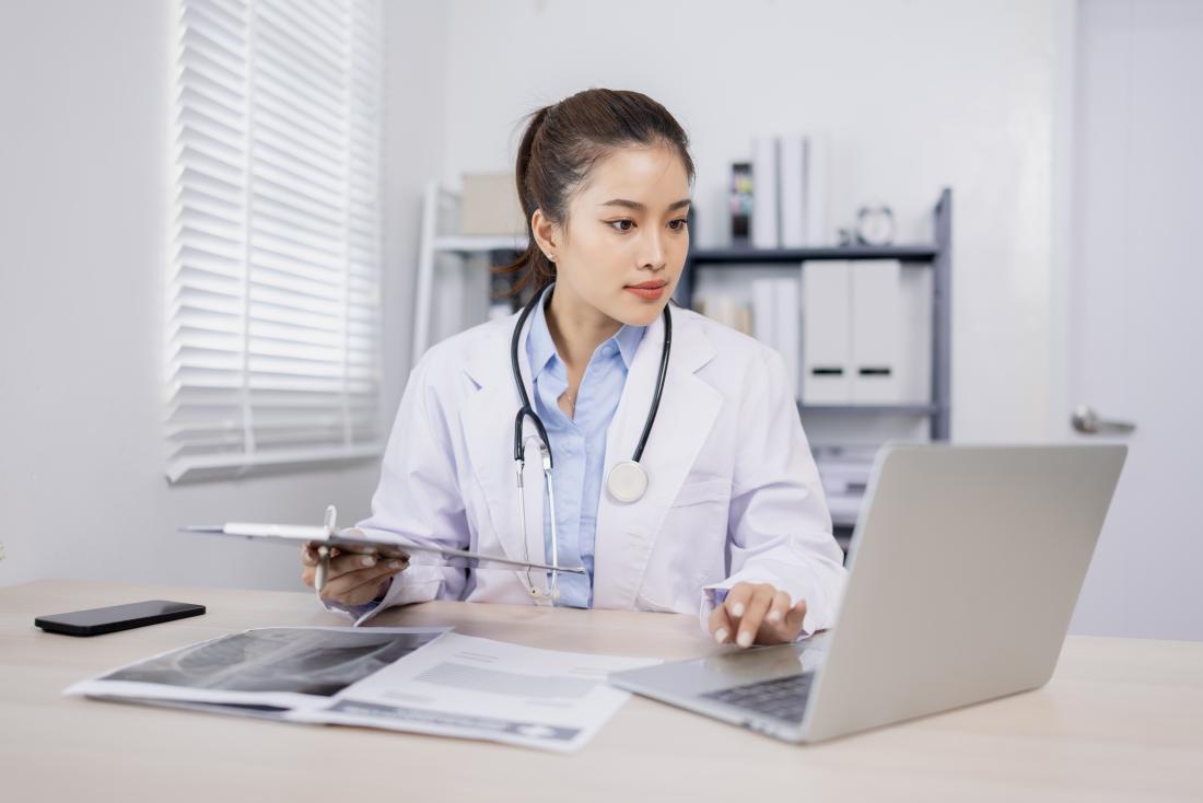female doctor in white coat sits at desk in front of laptop