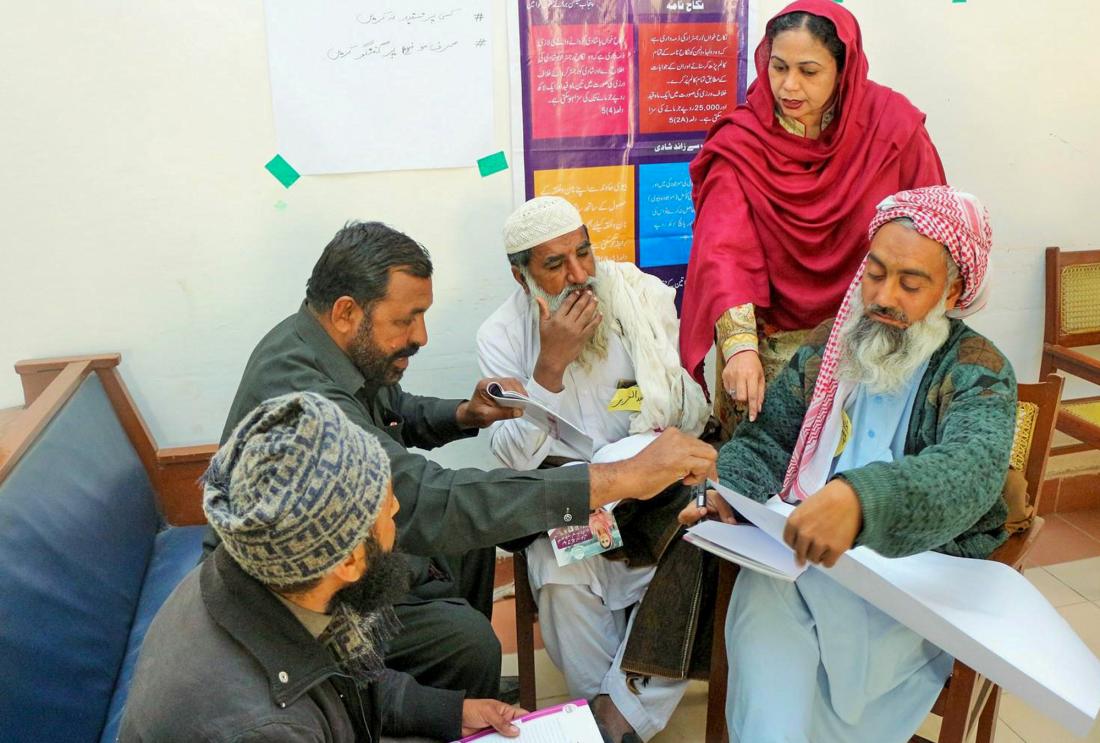 Woman stands over four men as they read papers. 