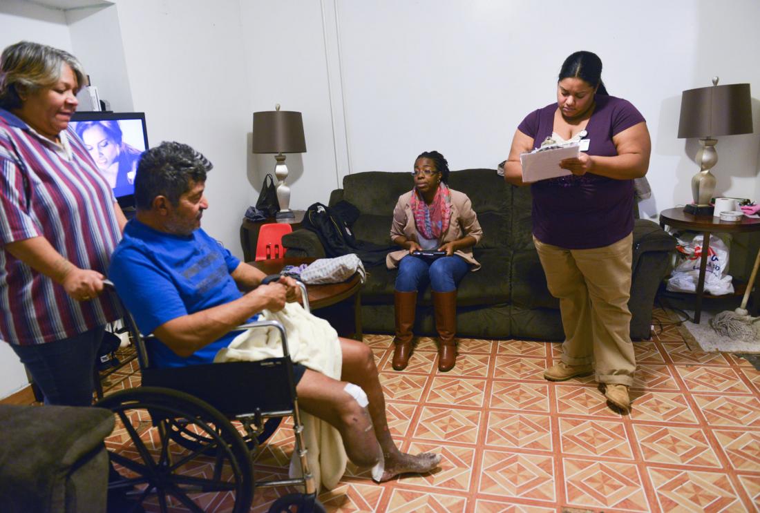 Two women with clipboards talk to man in wheelchair in living room