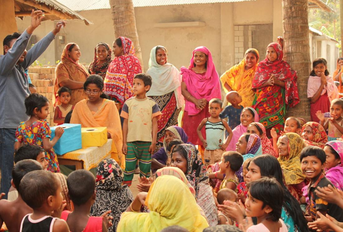 Crowd of women in bright saris watch man hold up token from cardboard box