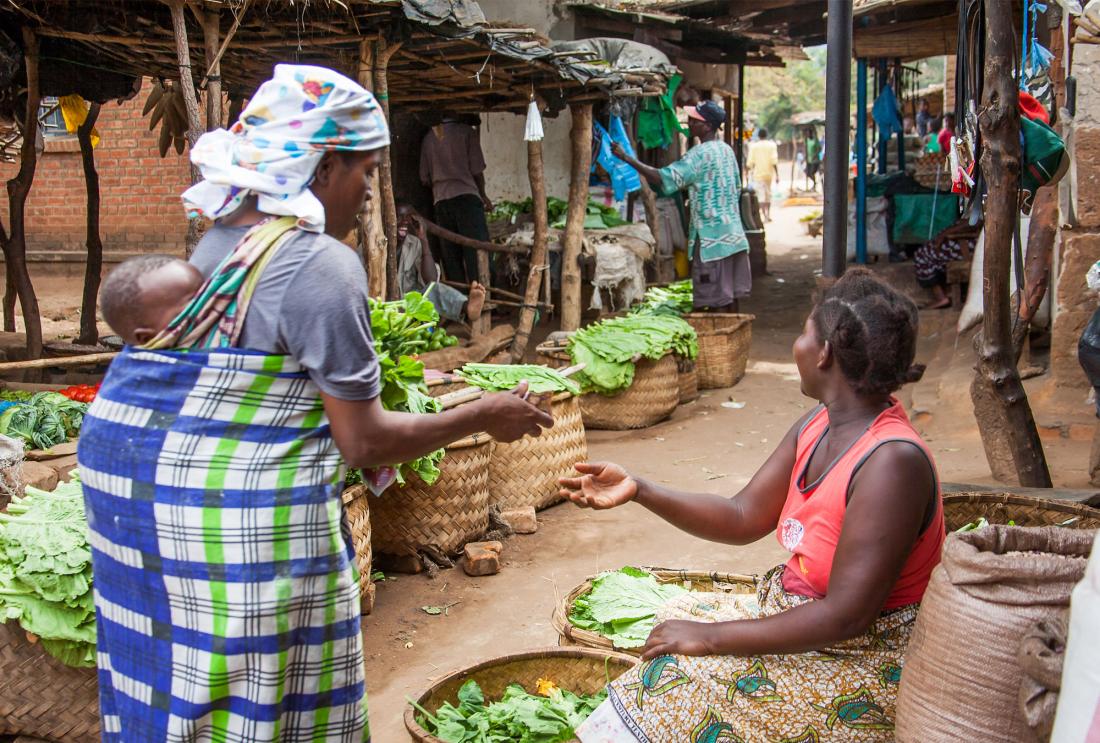 Woman carrying baby on her back pays for her greens at a rural market in Malawi