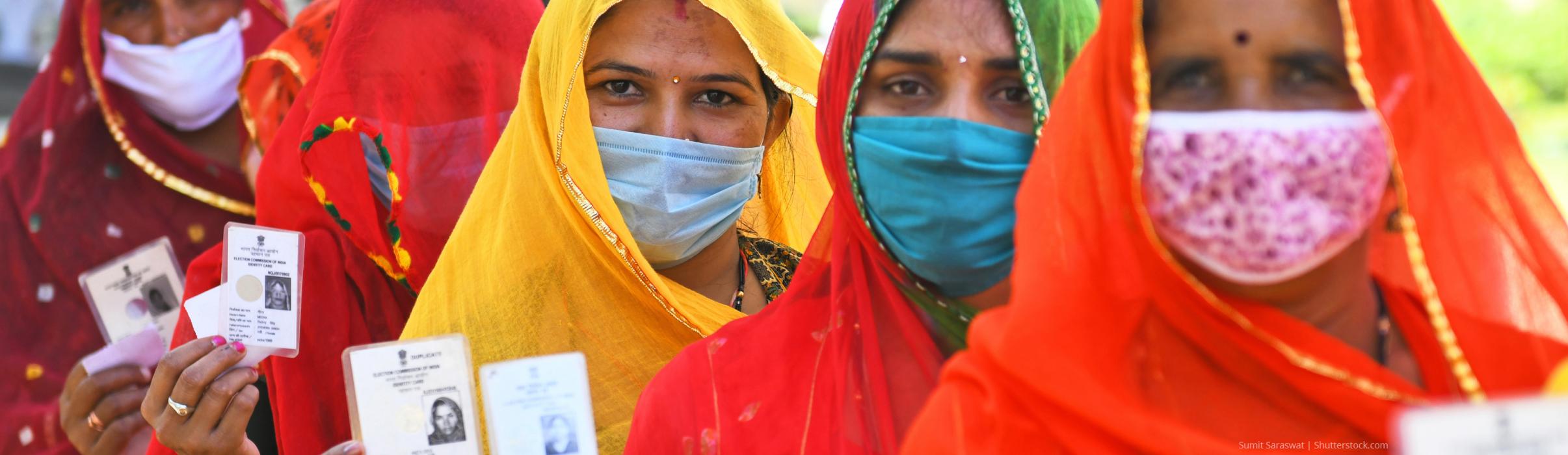 Women wearing face masks stand in a row holding ID cards while waiting to cast votes in Panchayati Raj elections during Covid-19