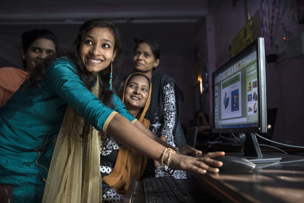 A group of four Indian woman stand in front of a computer and smile at something off-camera