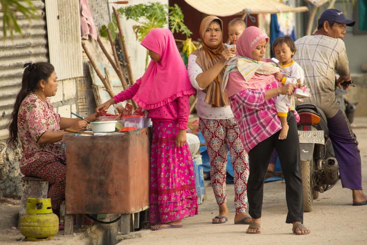Three women, two holding babies, buy food from a vendor seated at a table.