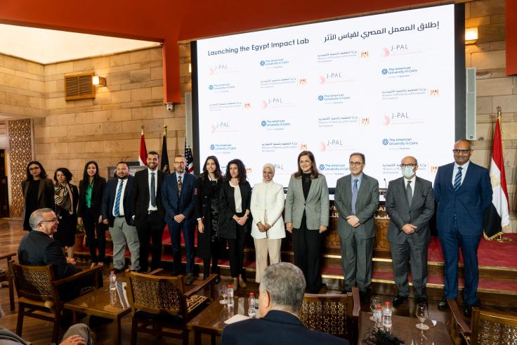 Thirteen people stand in a row in front of a screen with J-PAL, AUC, and MPED logos on it