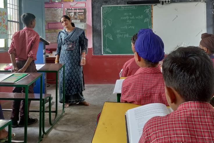 Teacher stands in front of classroom next to male standing student, while other students are seated at desks