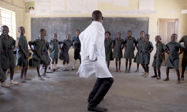 A group of children in a classroom encircle a person dancing in front of them.