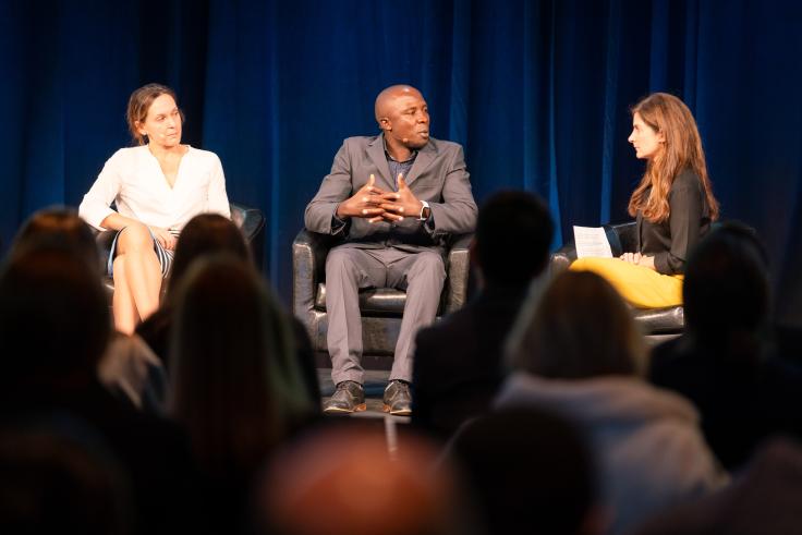 Two woman and one man are seated on a stage, in dialogue.