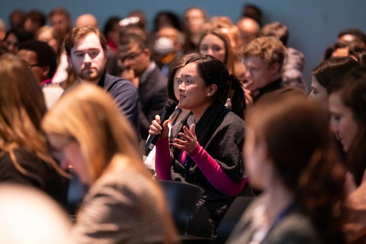 A woman seated in an audience holds a microphone during audience Q&A. 