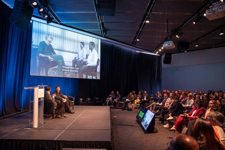 Audience members watch a video interview projected on a large screen above a stage.