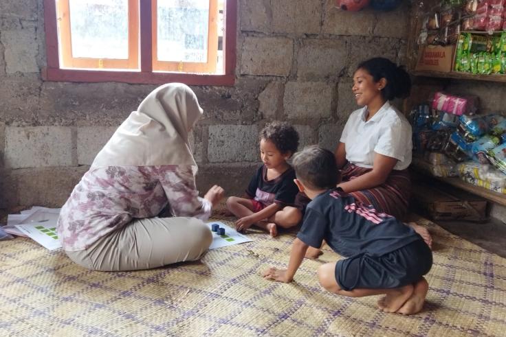 Facilitator sitting across from two children doing an activity with a caregiver smiling behind them