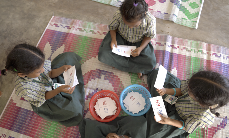 Three young girls in school uniforms sit cross-legged on colorful floor mats, engaged in a math activity using number flashcards. 