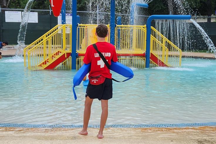 Teenage boy standing in shallow water wearing a lifeguard shirt and holding a rescue float