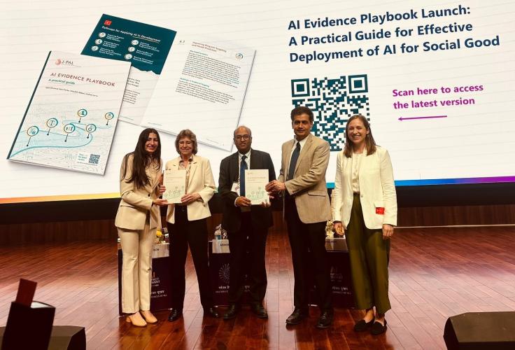 Five men and women stand together on a stage facing the audience. One man is displaying a book to the audience. The background reads, "Launch of the AI Evidence Playbook."