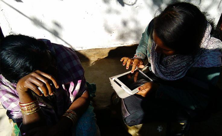 Two women sit together, one entering data into a tablet